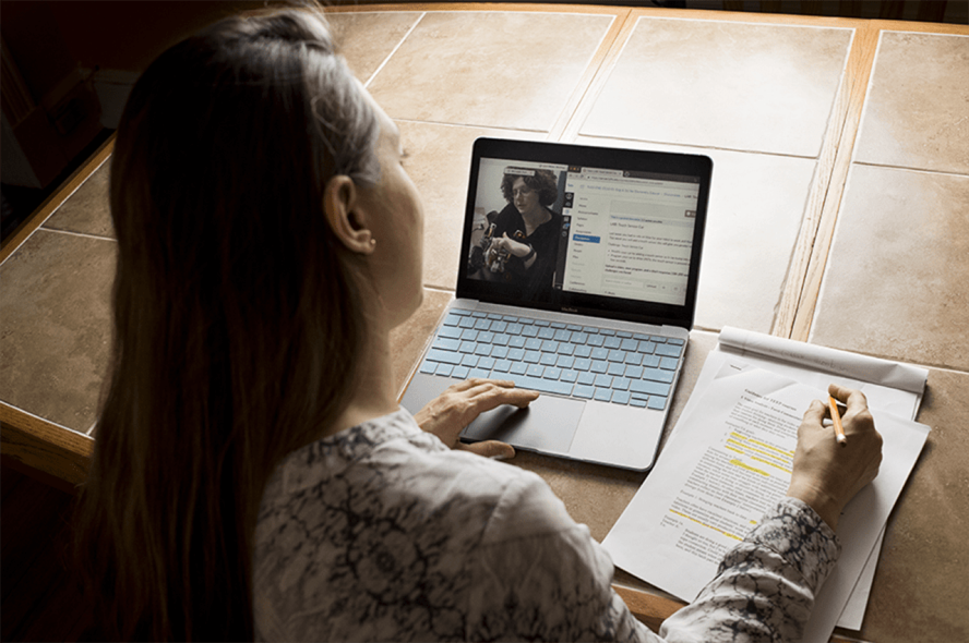 Woman sits at a counter watching a video on her laptop and taking notes by hand.